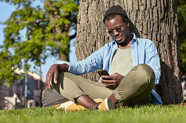 Young man sat in the sun on his phone. He is smiling lightly.