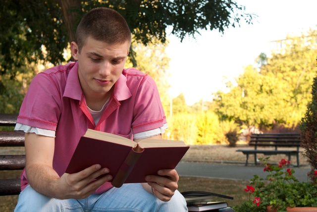 a young man sat on a bench reading a book outside