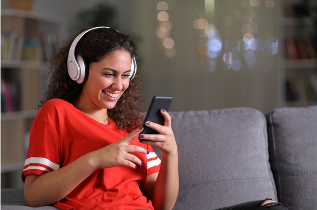 A young person sits on their sofa wearing a red t-shirt and headphones; they are looking at their phone and smiling