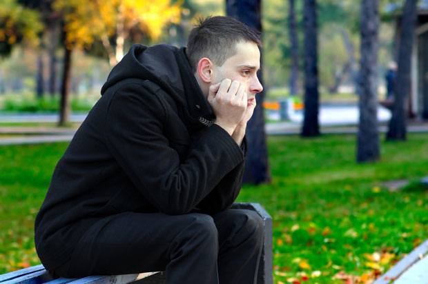 young man sits on bench in park looking upset.