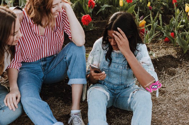 Three friends sitting down in a field looking at their mobiles and speaking to one another