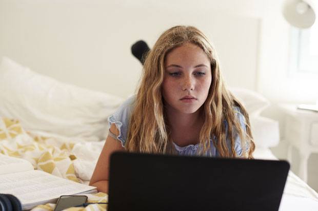 Girl lies on bed in her room looking at laptop with a book and phone by her side.