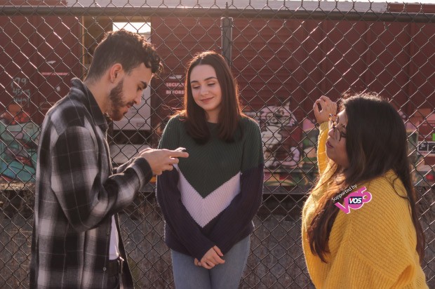 Three friends hanging out together . Far left a boy witha checkered shirt on his phone, middle girl leaning on a metal fence, left a girl in a yellow jumper with one hand on fence.