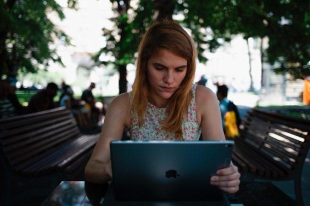 a young woman sits at a bench in a park and works on a laptop